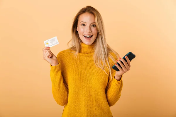 クレジット カード 携帯電話の使用を示す 黄色の背景に分離立ってセーターを着て興奮した若い女性の肖像画 — ストック写真
