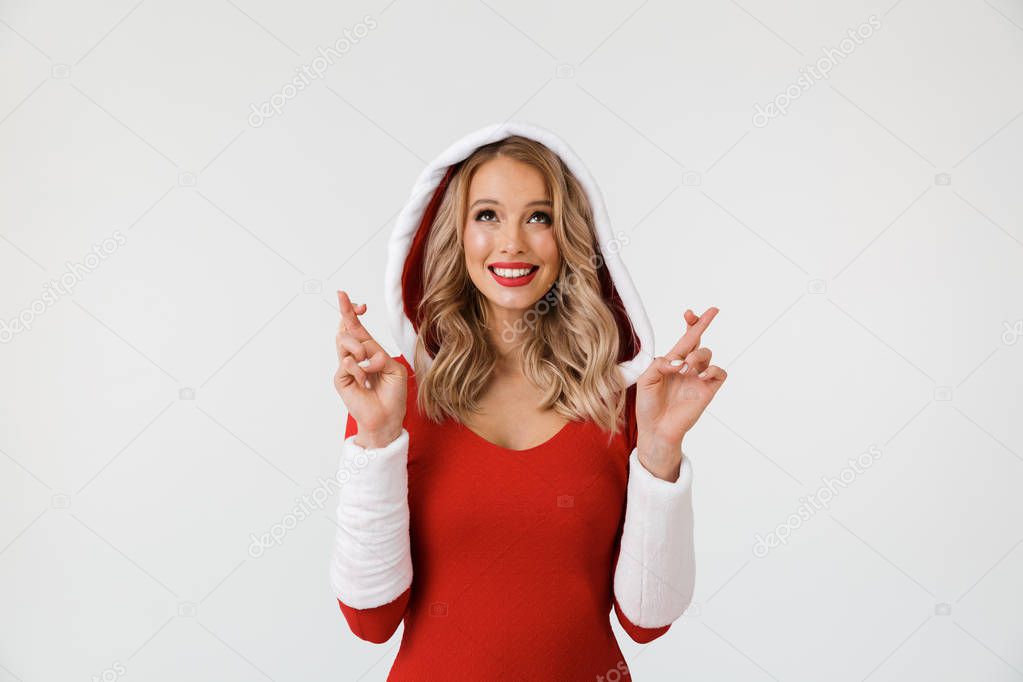Portrait of a happy blonde woman dressed in red New Year costume standing isolated over white background, holding fingers crossed for good luck