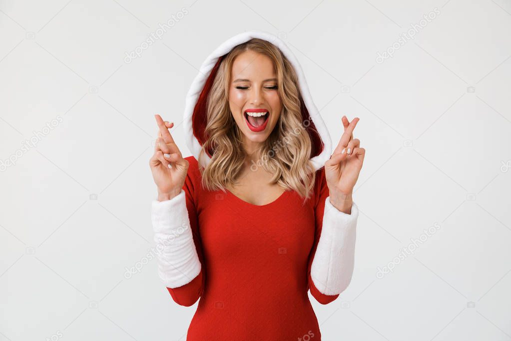 Portrait of a joyful blonde woman dressed in red New Year costume standing isolated over white background, holding fingers crossed for good luck