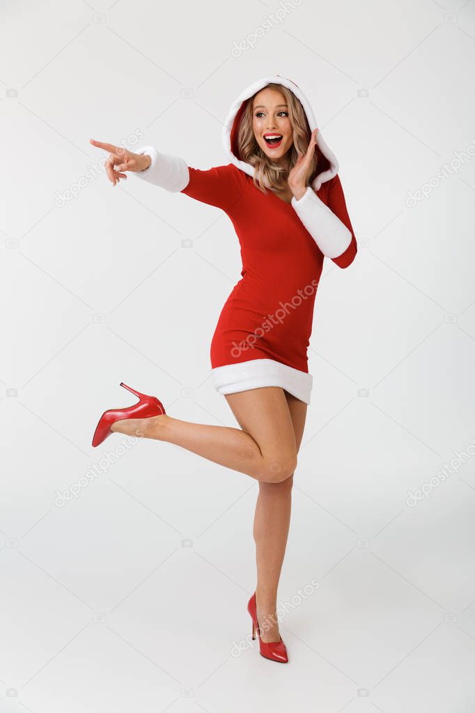 Full length portrait of a cheerful blonde woman dressed in red New Year costume standing isolated over white background, pointing finger at copy space
