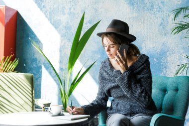 Üstünde hareket eden telefon, şık iç mekanlara söz güzel gülümseyen genç kadın kazak ve kafe masasında, sandalyede oturan şapka giymiş