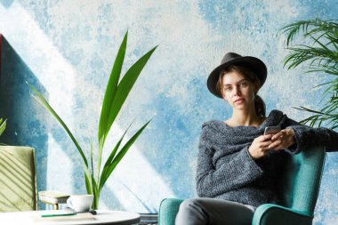 Güzel genç kadın kazak ve Cafe tablo, holding hareket eden telefon, şık iç mekanlara sandalyede oturan şapka giymiş