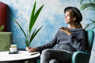 Güzel genç kadın kazak ve kafe masasında, sandalyede oturan şapka üstünde hareket eden telefon, şık iç mekanlara, notlar alarak konuşmaya giymiş