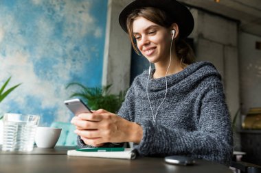 Kulaklık, şık iç mekanlara, Not almak ile müzik dinlemek güzel genç kadın kazak ve kafe masasında, sandalyede oturan şapka giymiş