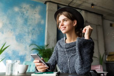 Kulaklık, şık iç mekanlara, Not almak ile müzik dinlemek güzel neşeli genç kadın kazak ve kafe masasında, sandalyede oturan şapka giymiş