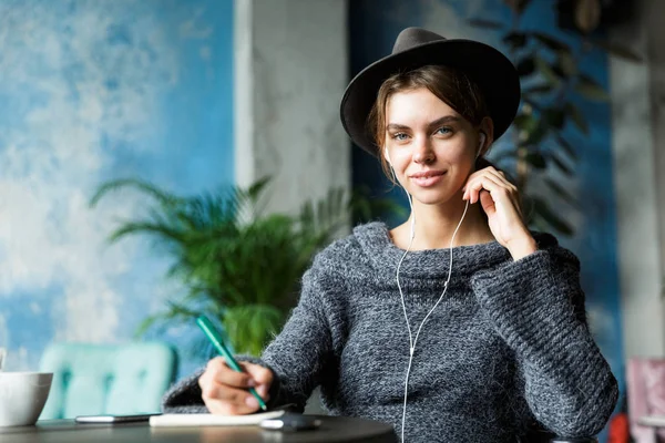 美丽的微笑的年轻女子穿着毛衣和帽子坐在椅子上的咖啡馆桌子 听音乐与耳机 时尚的内饰 做笔记 — 图库照片