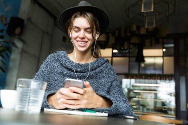 İstimal hareket eden telefon, şık iç mekanlara kulaklık ile müzik dinlemek güzel genç kadın kazak ve kafe masasında, sandalyede oturan şapka giymiş
