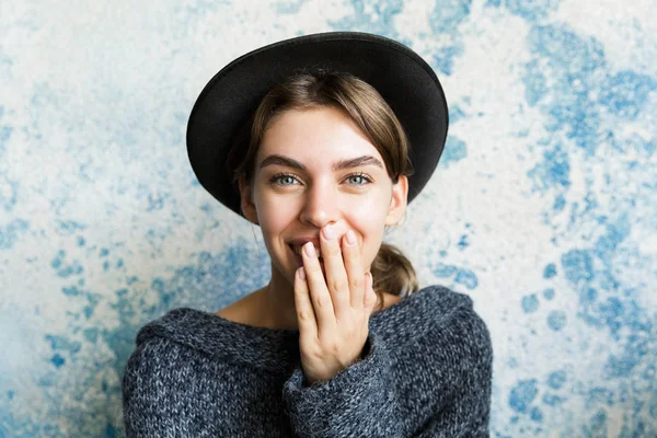 クローズ アップ カメラ 見て笑って カバー口青い壁の背景の上のセーターと帽子に身を包んだ若い女性の肖像画 — ストック写真