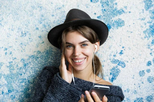 Close up portrait of a cute young woman dressed in hat and sweater listening to music with eaphones, holding mobile phone over blue wall background