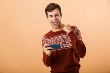 Portre cep telefonunda oyun bej renkli arka plan üzerinde izole bir heyecanlı genç adam ayakta kutluyor