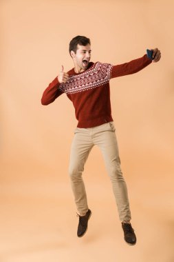 Tam uzunlukta portre selfie cep telefonu ile alarak bej renkli arka plan üzerinde izole atlama neşeli genç bir adam