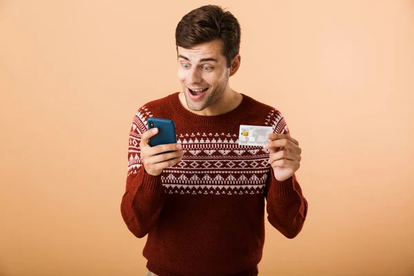 肖像一个开朗的年轻人穿着毛衣站在米色背景孤立 拿着手机 展示信用卡 — 图库照片