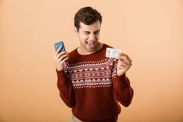 肖像一个兴奋的年轻人穿着毛衣站在米色背景孤立 拿着手机 展示信用卡 — 图库照片