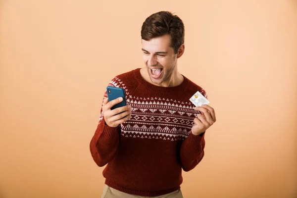 肖像一个开朗的年轻人穿着毛衣站在米色背景孤立 拿着手机 展示信用卡 — 图库照片