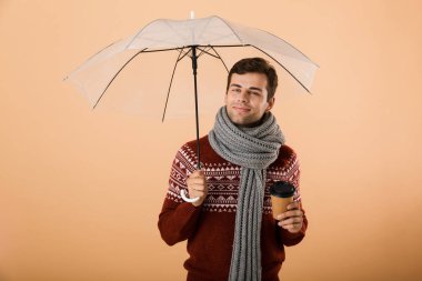 Kazak ve kahve fincanı holding şemsiyesi altında duran bej renkli arka plan üzerinde izole eşarp portre mutlu bir genç adam giymiş