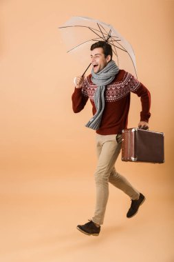 Kazak ve eşarp çanta taşıyan bir şemsiye altında yürüyüş bej renkli arka plan üzerinde izole tam uzunlukta portre heyecanlı bir genç adam giymiş