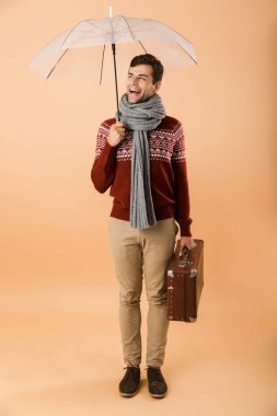 Kazak ve eşarp çanta taşıyan bir şemsiye altında duran bej renkli arka plan üzerinde izole tam uzunlukta portre genç gülümseyen bir adam giymiş