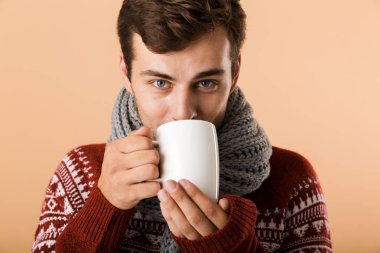 Kazak ve bej renkli arka plan üzerinde izole bir fincan çay içme eşarp giymiş memnun genç adam, yakın çekim