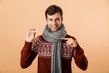 Kazak ve eşarp giymiş mutlu bir genç adam portresi termometre gösterilen, parmak işaret bej renkli arka plan üzerinde izole