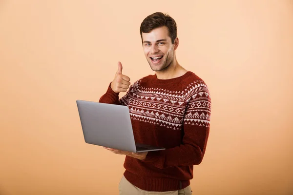 分離親指を現して ラップトップ コンピューターを使用して ベージュの背景の上に立っているセーターを着て陽気な若い男の肖像画 — ストック写真