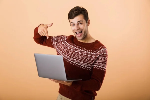 ベージュ色の背景 ラップトップ コンピューターを使用して 指を指して立って分離セーターに身を包んだ陽気な若い男の肖像画 — ストック写真