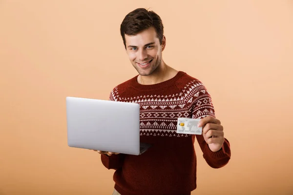 一个开朗的年轻人 穿着米色背景上的毛衣 拿着笔记本电脑 展示信用卡 — 图库照片