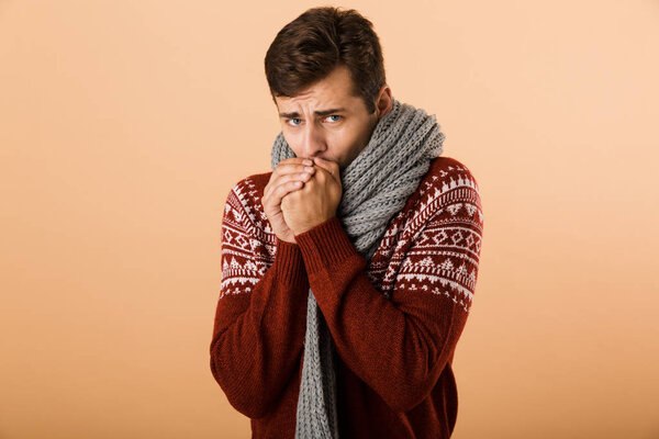 Portrait a young man dressed in sweater and scarf isolated over beige background, warming up arms