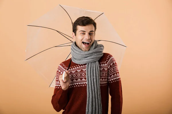 幸せな若い男の肖像画を着たセーター マフラーの傘の下に立って ベージュの背景の上分離 — ストック写真