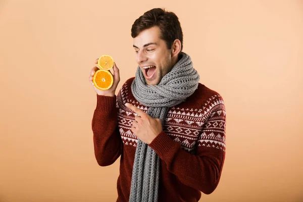 Portrait a happy man dressed in sweater and scarf isolated over beige background, holding sliced oranges