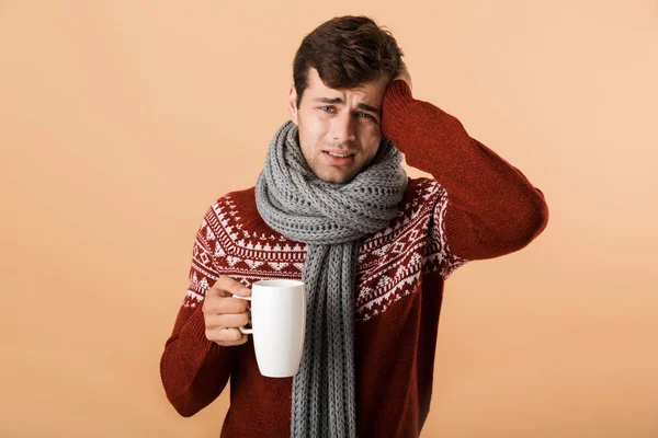 セーターとスカーフ頭痛 ベージュの背景の上分離された茶のカップを保持している服を着て悲しい若い男の肖像 — ストック写真