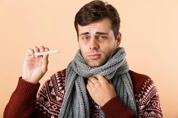 一个悲伤的生病的年轻人的肖像 穿着毛衣和围巾隔离在米色的背景 显示温度计 — 图库照片