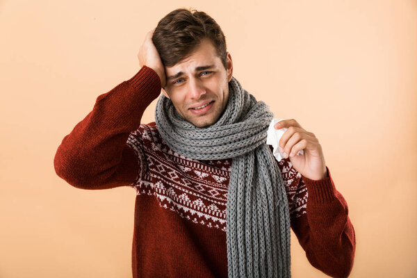 Portrait of a sick young man dressed in sweater and scarf isolated over beige background, holding a tissue