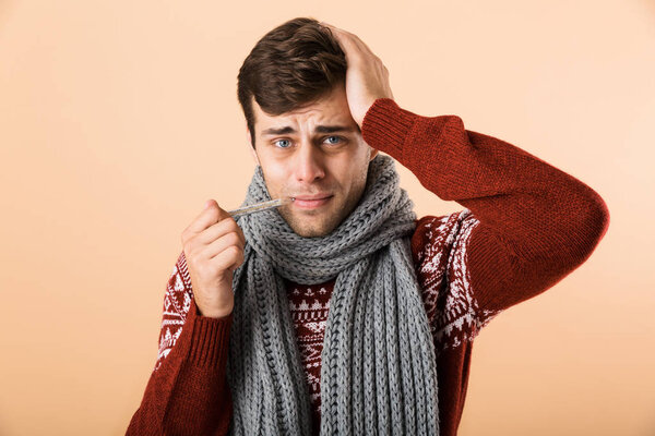 Portrait of a sick young man dressed in sweater and scarf isolated over beige background, showing thermometer