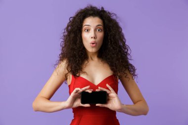 Görüntü siyah smartphone menekşe arka plan üzerinde izole ayakta tutan memnun kadın 20'li yaşlarda giyen kırmızı elbise