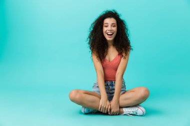 Bacaklar ile katta otururken genç kadın 20'li yaşlarda casual giyim giyen gülen görüntü izole mavi arka plan üzerinde çarpı işareti