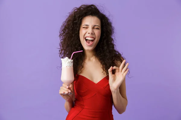 一个高兴的女人的肖像与深色卷发穿着红色礼服隔离在紫罗兰色背景 喝奶昔从玻璃 显示确定 — 图库照片