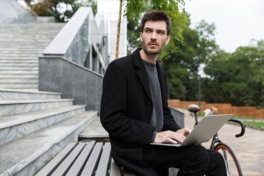 Açık havada dizüstü bilgisayar kullanan oturan yakışıklı genç iş adamı görüntüsü.