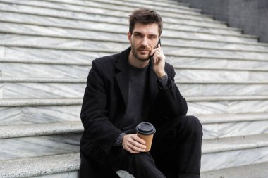 Açık havada kahve içme cep telefonuyla konuşurken oturan yakışıklı genç iş adamı görüntüsü.