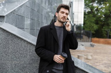 Açık havada kahve içme cep telefonuyla konuşurken oturan yakışıklı genç iş adamı görüntüsü.