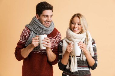 Kazak ve birlikte ayakta eşarp genç gülümseyen bir çift portresi bardak çay ile tutarak bej renkli arka plan üzerinde izole