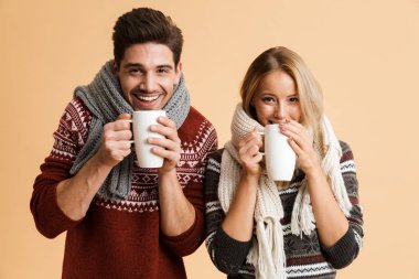Kazak ve birlikte ayakta eşarp genç gülümseyen bir çift portresi bardak çay ile tutarak bej renkli arka plan üzerinde izole