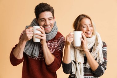 Bardak çay ile tutarak bej renkli arka plan üzerinde memnun genç bir çift kazak ve eşarplar birlikte ayakta giymiş portresi izole