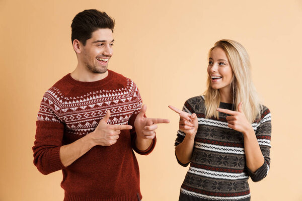 Portrait of a happy young couple dressed in sweaters standing together isolated over beige background, pointing at each other