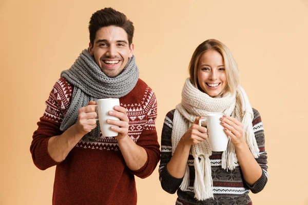 お茶とカップを保持しているベージュの背景の上分離したセーターと一緒に立っているスカーフを着て幸せな若いカップルの肖像画 — ストック写真