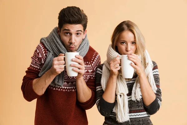お茶とカップを保持しているベージュの背景の上分離したセーターとマフラー一緒に立って服を着た素敵な若いカップルの肖像画 — ストック写真