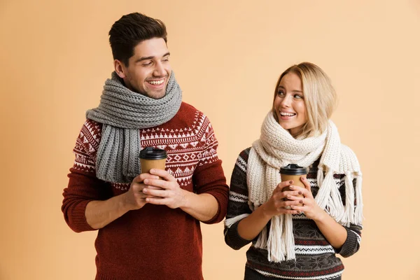 コーヒー カップを保持しているベージュの背景の上分離したセーターと一緒に立っているスカーフを着て満足している若いカップルの肖像画 — ストック写真