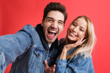 Portre heyecanlı bir genç çiftin birlikte ayakta kot ceketler içinde giyinmiş bir selfie alarak, kırmızı arka plan üzerinde izole