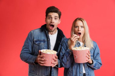 Portre şok bir genç çiftin birlikte ayakta kot ceketler içinde giyinmiş film izlerken, patlamış mısır yeme kırmızı arka plan üzerinde izole