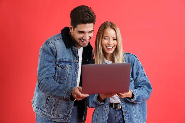 一个穿着粗斜纹棉布夹克的年轻夫妇的肖像站在一起 孤立在红色的背景 看着笔记本电脑 — 图库照片