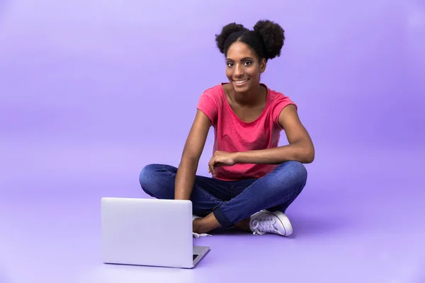 照片年轻的非洲裔美国妇女微笑着 使用银色笔记本电脑 而坐在地板上与腿交叉交叉在紫罗兰色背景 — 图库照片